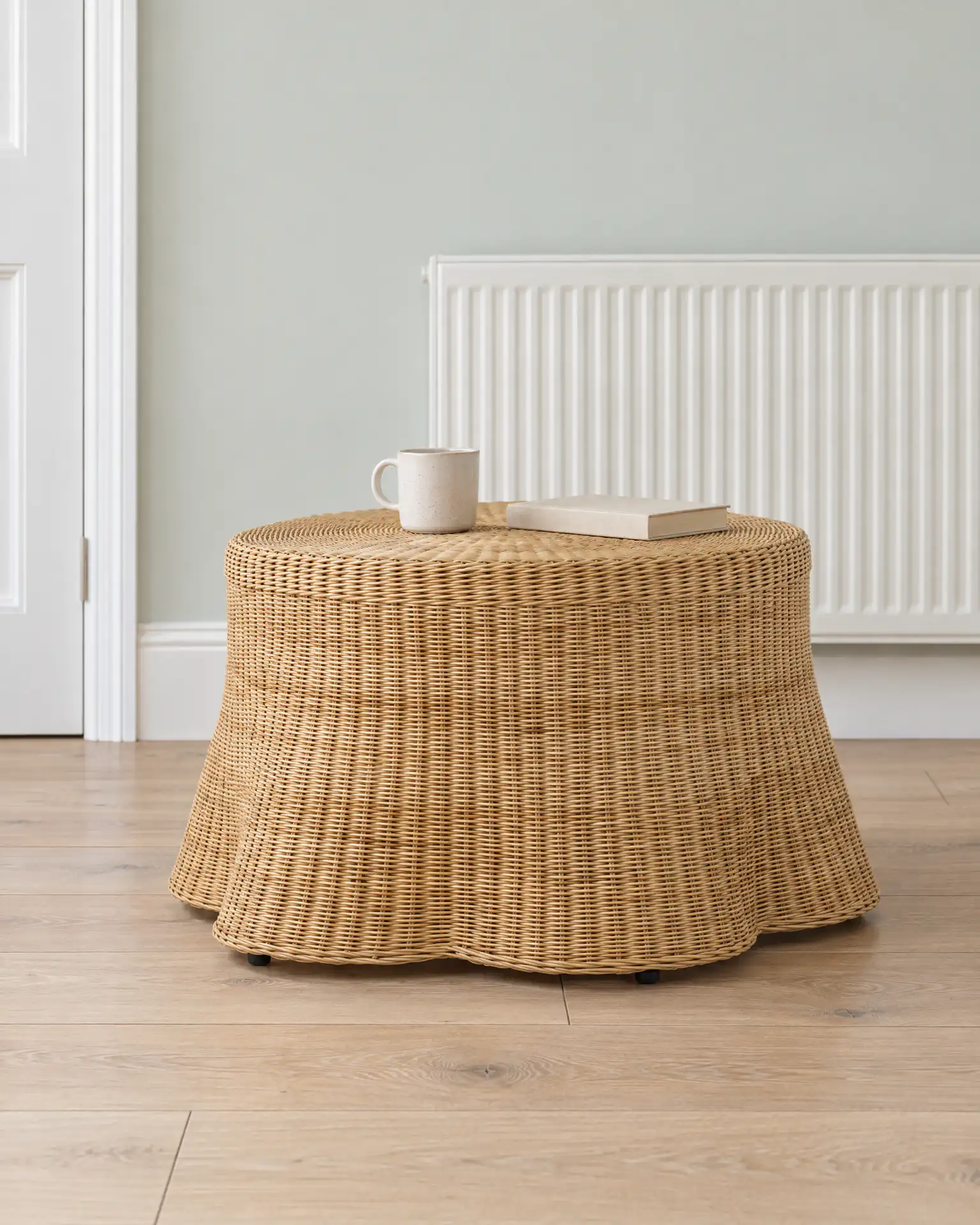 The round rattan coffee table with its scalloped hem in front of a white radiator, a cup and a book on top.
