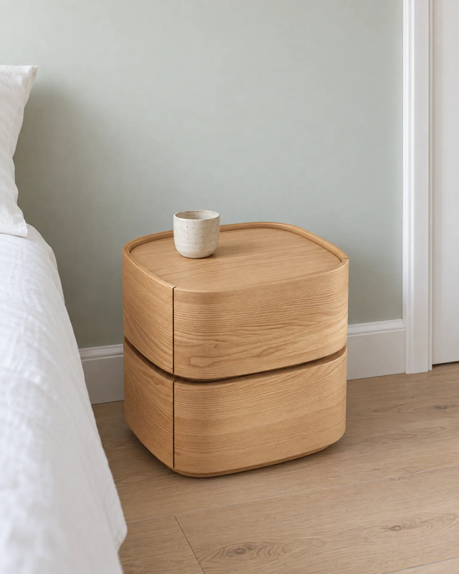 The curved wood bedside table with two drawers against a pale green wall, beside the corner of a bed, a small cup on top.