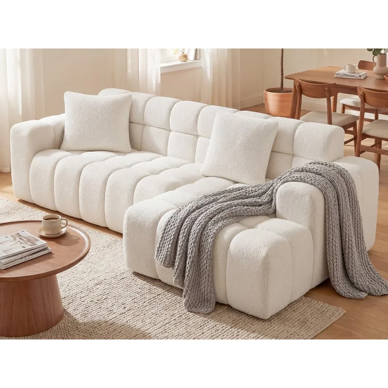 The white tufted corner sofa in a bright living room, a grey knitted throw over the chaise, a round wooden coffee table in front